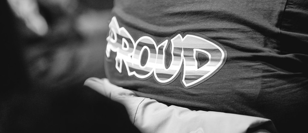 Black and white image of a woman's back sitting in a chair with the word proud on the back of her t-shirt